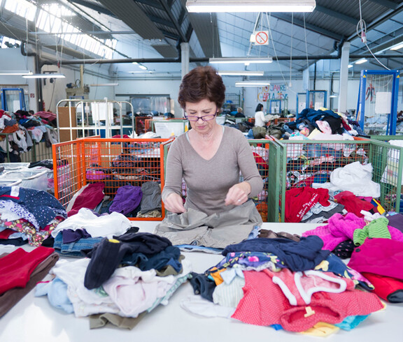 Eine carla Mitarbeiterin mittleren Alters mit rot gefärbten, kurzen Haaren und einer Brille steht hinter einem großen Tisch und sortiert Kleidung. 