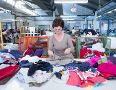 Eine carla Mitarbeiterin mittleren Alters mit rot gefärbten, kurzen Haaren und einer Brille steht hinter einem großen Tisch und sortiert Kleidung. 