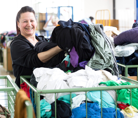 Eine carla Mitarbeiterin mittleren Alters mit langen schwarzen Haaren sortiert fröhlich und heiter Kleidung aus einem grünen Schiebewagen. 