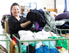 Eine carla Mitarbeiterin mittleren Alters mit langen schwarzen Haaren sortiert fröhlich und heiter Kleidung aus einem grünen Schiebewagen. 